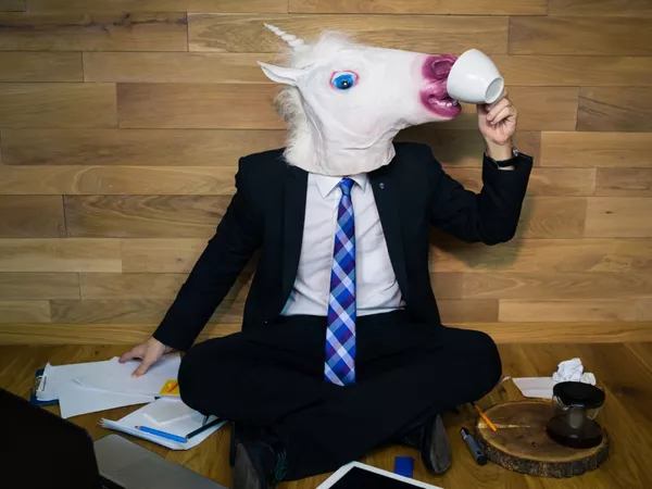 Man seating on the floor wearing a suit and an unicorn head custom. He is trying to drink from a white cup and has laptop and some paper on the floor next to him. 