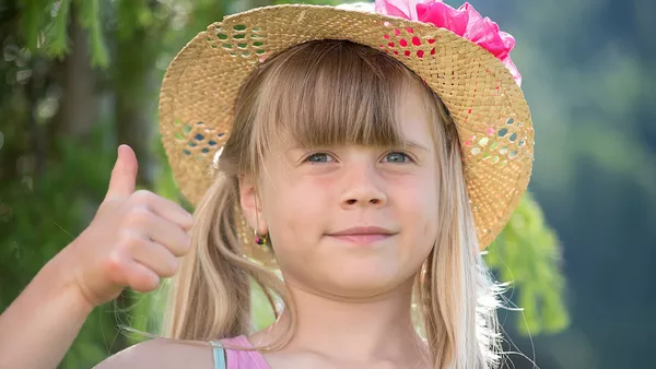A young child with blonde hair and a summer hat giving a blank expression thumbs up to the camera.