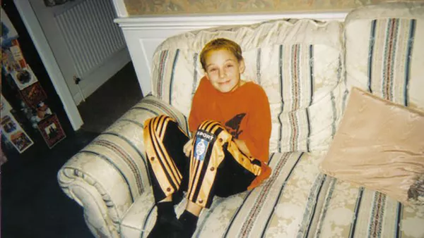 Darren as a young man (circa 10 years old) sat on a white sofa with stripes, wearing an orange t-shirt and black and orange tracksuit bottoms.