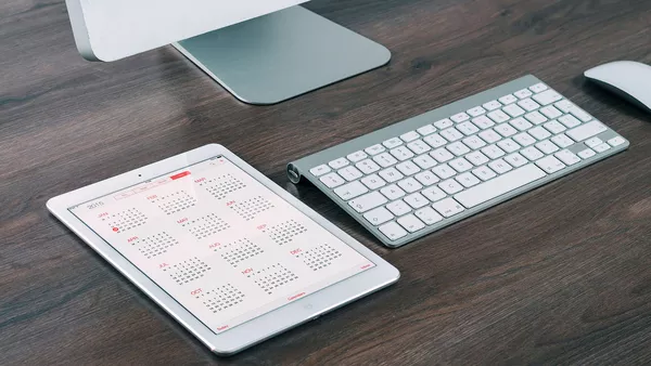 A calendar app open on an ipad laying on a desk next to an imac, keyboard and mouse.