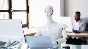 A robot at an office desk working with a laptop and another man working in desk behind him using a computer. 