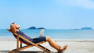A man in shorts and t-shirt lying back in a deckchair on a white sandy beach with a laptop on his lap.